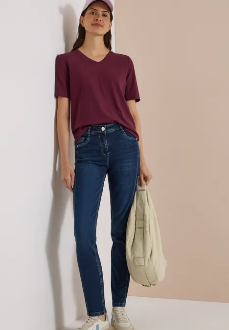 Jeune femme en T-shirt violet, jean et casquette, appuyée contre un mur beige, tient un sac en tissu clair.