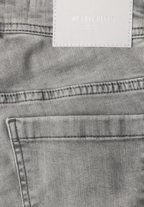 Close-up van de achterkant van een grijze spijkerbroek met achterzak, lussen en label “WE LOVE DENIM”.