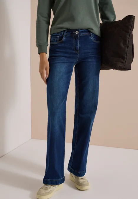 Person trägt dunkelblaue, ausgestellte Jeans, grünes Langarmshirt und hält ein dunkles Kissen.