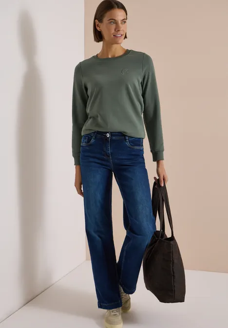 Frau in grünem Pullover, blauen Jeans und beigen Schuhen trägt schwarze Tasche vor beiger Wand.