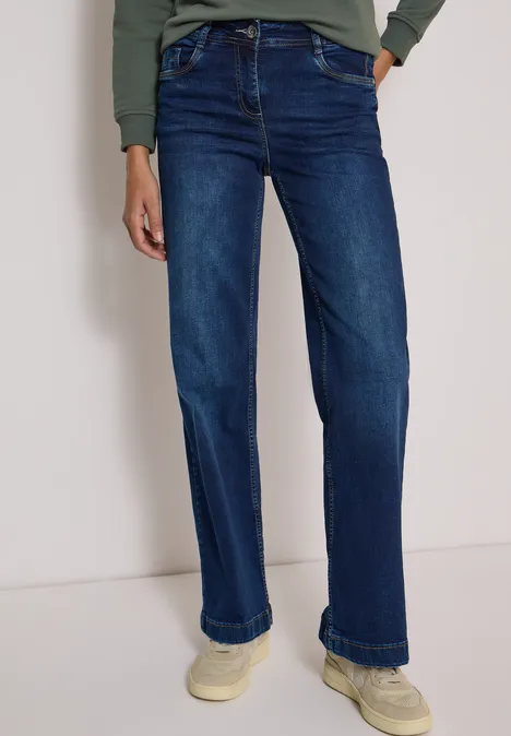 Person in dunkelblauen, geraden Jeans und hellen Sneakers vor neutralem Hintergrund.