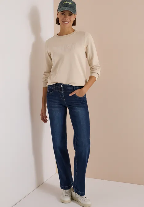 Eine Frau in Jeans, beigem Langarmshirt, weißen Sneakern und Cap steht lächelnd an einer Wand.