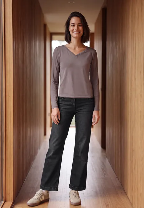 Een vrouw staat glimlachend in een houten gang, draagt een donkere jeans en een taupe longsleeve.
