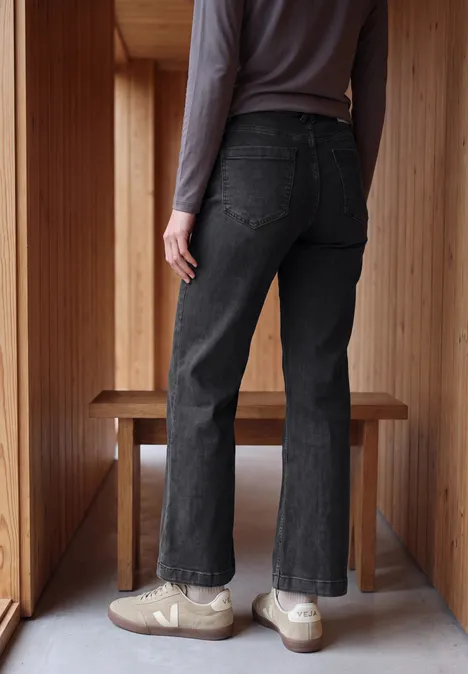 Iemand van achteren met zwarte jeans en beige sneakers, staand in een lichte houten ruimte.