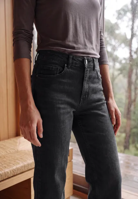 Vrouw in een grijs shirt met lange mouwen en zwarte high-waist jeans, staand voor raam met boszicht.