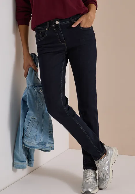 Model lehnt an Wand, trägt dunkelblaue Jeans, silberne Sneakers und hält eine hellblaue Jeansjacke.