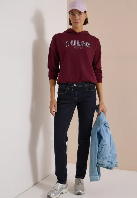 Frau in burgunderrotem Pullover, Jeans und Cap, hält eine Jeansjacke, trägt silberne Sneaker im Minimal-Setting.