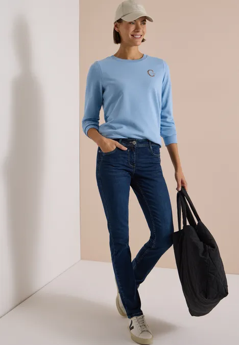 Frau mit hellblauem Shirt, dunklen Jeans, weißer Kappe und großer schwarzer Tasche lächelt entspannt.