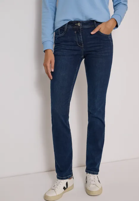 Frau in dunkelblauen Slim-Fit-Jeans, hellblauem Oberteil und weißen Sneakern vor neutraler Wand.