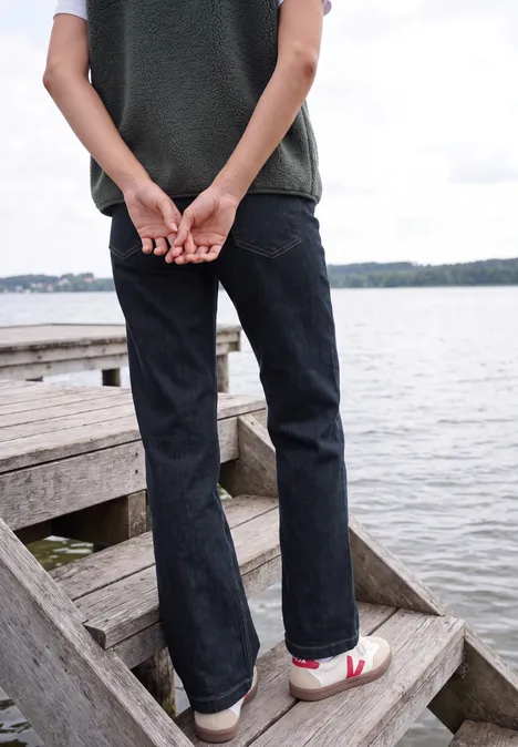 Person steht in Jeans und Sneakers auf einer Holztreppe am See, Hände hinter dem Rücken verschränkt.