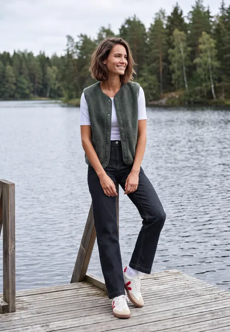 Frau lehnt lächelnd an einem Steg am See, trägt Weste, T-Shirt, Jeans und Sneaker.