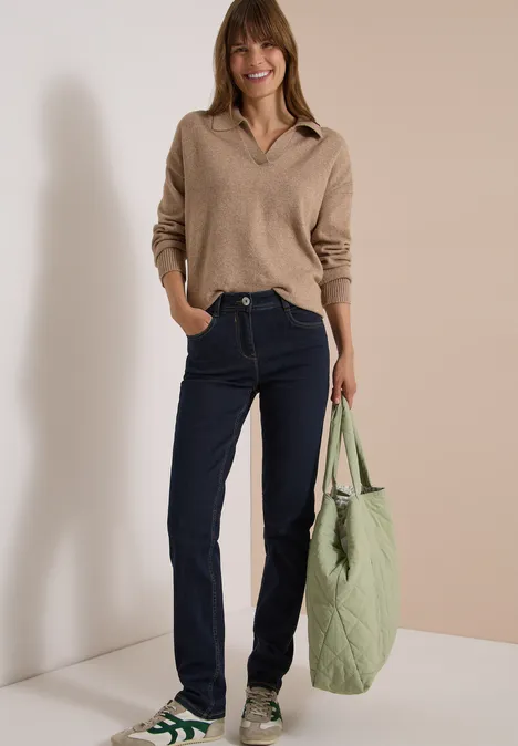 High Waist Slim Leg Jeans Slim Fit rinsed wash Frau in beigem Pullover, Jeans und grünen Sneakers, trägt eine große gesteppte grüne Tasche.