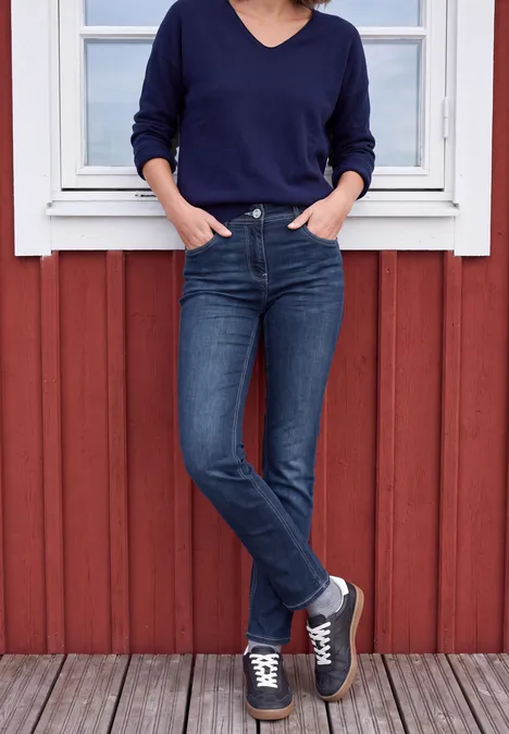 Vrouw in donkerblauwe V-trui en skinny jeans, handen in zakken, staand voor een rood houten gevel.