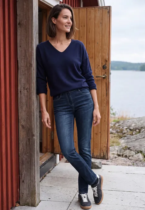 Een vrouw in jeans en blauwe trui, glimlachend tegen een houten gevel bij een meer.