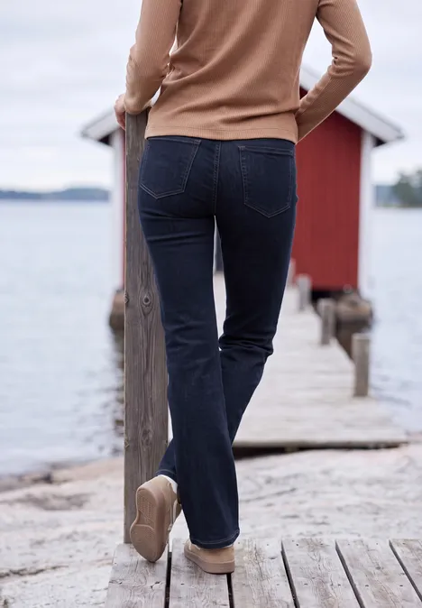High Waist Bootcut Jeans im Slim Fit rinsed wash Frau in Jeans und Pullover geht über Steg zum roten Bootshaus am See.