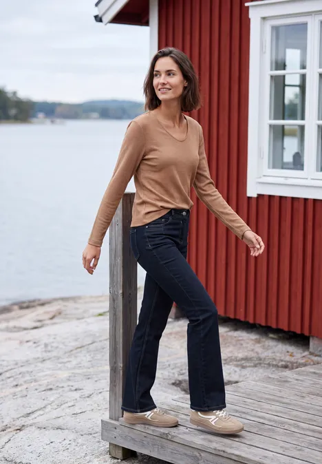 High Waist Bootcut Jeans im Slim Fit rinsed wash Frau in braunem Pullover und dunkelblauen Jeans spaziert lächelnd auf Holzsteg vor roter Hütte am Wasser.