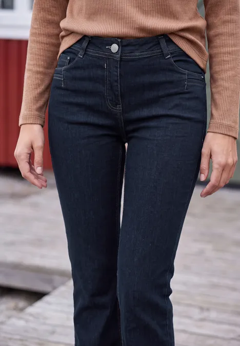 High Waist Bootcut Jeans im Slim Fit rinsed wash Eine Person trägt einen beigefarbenen Pullover und dunkelblaue Slim Jeans, stehend auf Holzplanken.
