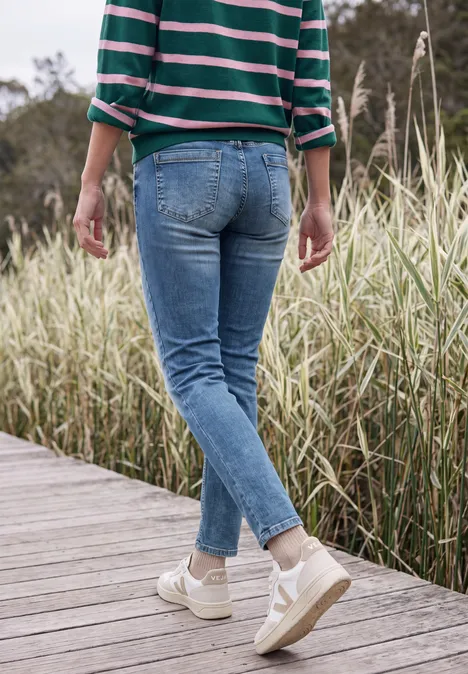 Mid Waist Slim Leg Jeans im Casual Fit mid blue wash Person läuft auf Holzsteg zwischen hohen Gräsern, trägt blaue Jeans und sandfarbene Sneakers.