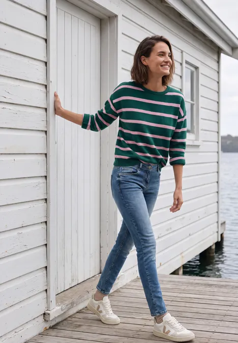Mid Waist Slim Leg Jeans im Casual Fit mid blue wash Frau in Jeans und gestreiftem Pullover spaziert lachend an einer weißen Strandhütte am Wasser entlang.