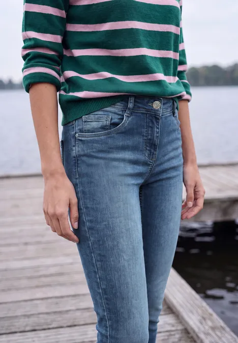 Mid Waist Slim Leg Jeans im Casual Fit mid blue wash Person steht am Wasserpier, trägt grün-weiß-rosa gestreiftes Oberteil und blaue Jeans.