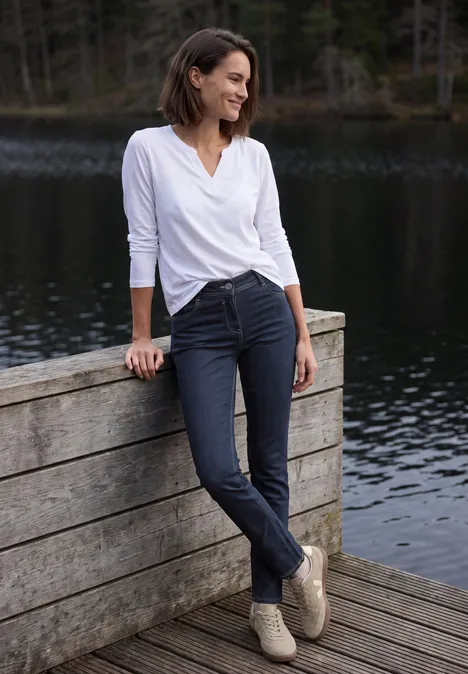 Mid Waist Straight Leg Jeans im Casual Fit rinsed wash Eine Frau lehnt entspannt an einem Holzgeländer am See, trägt weiße Bluse, dunkle Hose und lächelt.