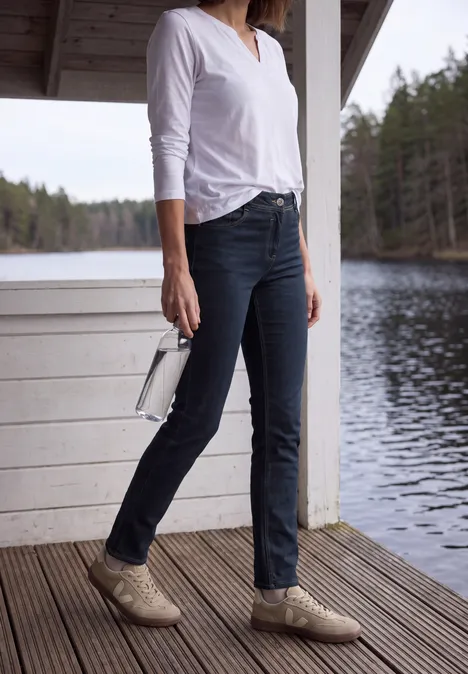 Mid Waist Straight Leg Jeans im Casual Fit rinsed wash Frau in weißem Shirt und dunklen Jeans, mit beigen Schuhen und weißer Tasche, steht auf Holzdiele am See.