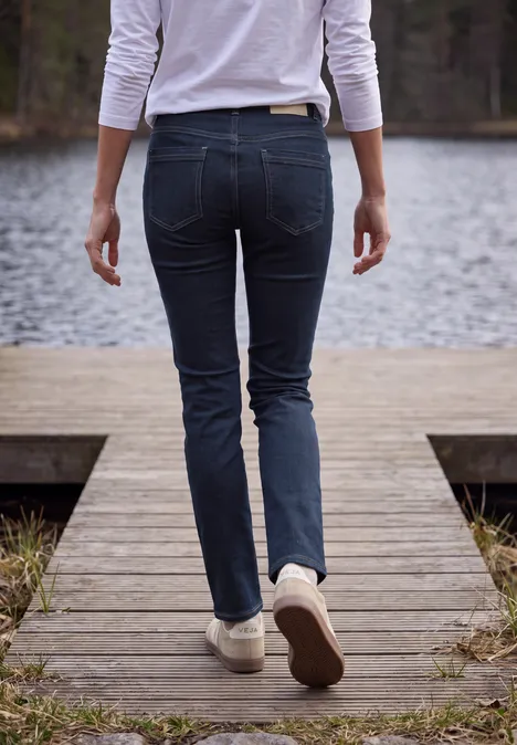 Mid Waist Straight Leg Jeans im Casual Fit rinsed wash Person in engen Jeans und weißen Sneakern geht auf einem Holzsteg am See entlang.