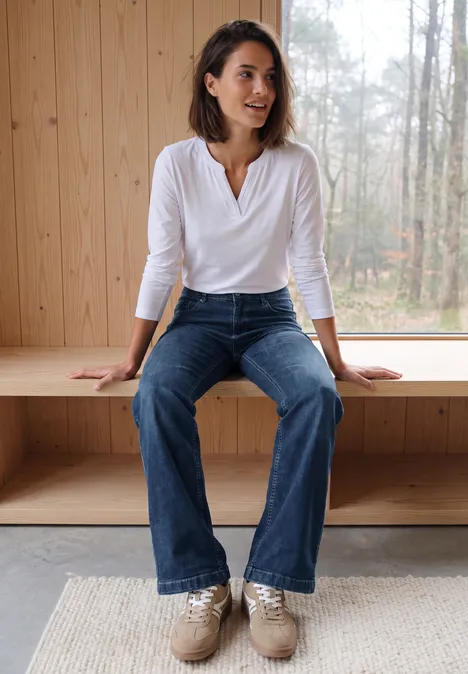 Wide Leg Jeans mit Gallonstreifen mid blue used wash Frau in weißem Shirt und Schlagjeans sitzt lächelnd auf einer Fensterbank vor einer Holzwand, Natur draußen.