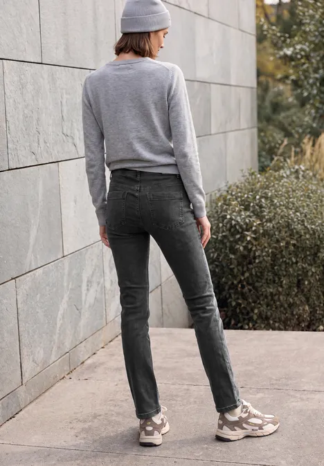Eine Person steht vor einer Steinwand in grauer Kleidung mit Jeans, Pullover, Mütze und Sneakern.