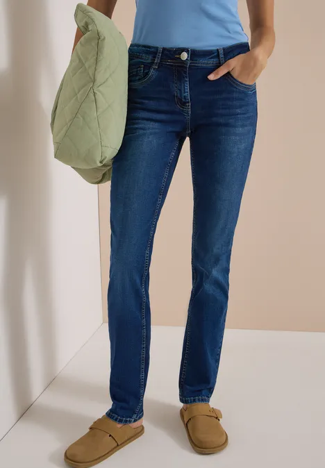 Une personne porte un jean bleu, un haut bleu clair et des chaussons marron, tenant un sac matelassé vert.