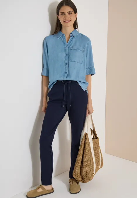 Femme en chemise bleue et pantalon sombre, tenant un grand sac en paille devant un mur clair.