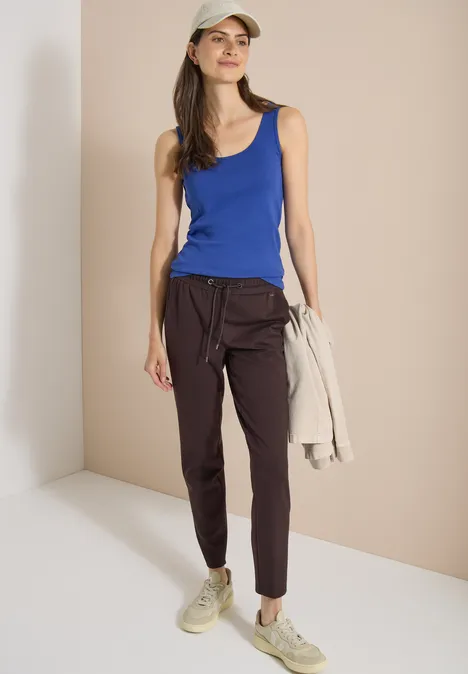 Een vrouw in een blauw tanktop, donkere broek en sneakers houdt een licht jasje vast.