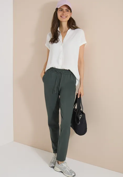 Vrouw in witte blouse en donker joggerbroek met aantrekkoord, witte sneakers en zwarte tas voor een beige muur.