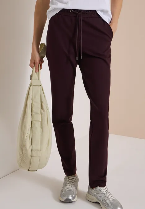 Une personne porte un chemisier blanc, un pantalon bordeaux, des baskets grises et un sac beige.