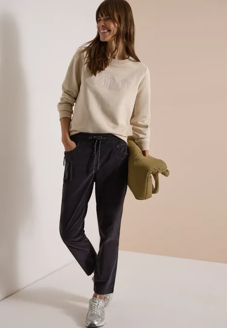 Femme en sweat beige, pantalon jog noir, baskets argent, tenant une veste kaki devant un mur clair.