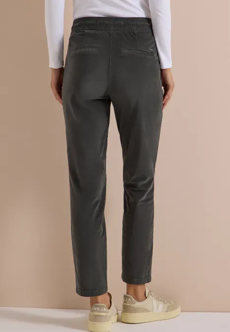 Vue arrière d’une personne portant un pantalon gris foncé droit et des baskets claires sur fond beige.
