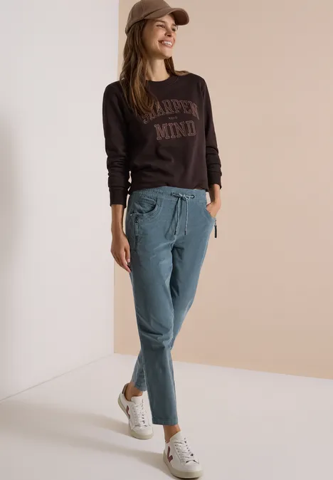 Femme en casquette avec sweat marron, pantalon bleu clair et baskets blanches, posant décontractée.