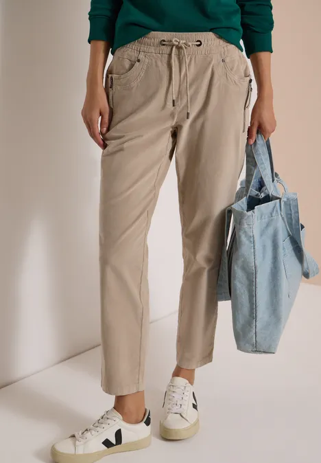 Pantalon beige à plis, baskets blanches et sac en tissu bleu tenu à la main.