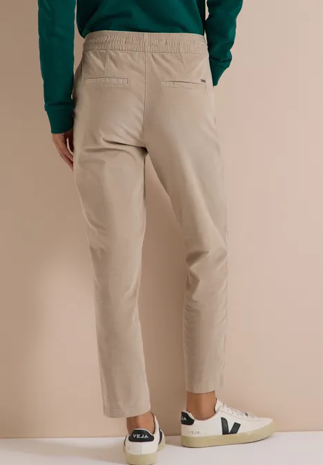 Vue arrière d'une personne portant un pantalon beige et des baskets blanches à logo V sur fond neutre.