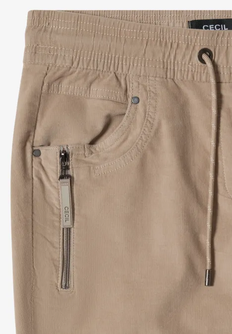 Pantalon beige en velours côtelé, taille élastique à cordon, poche zippée et surpiqûres.