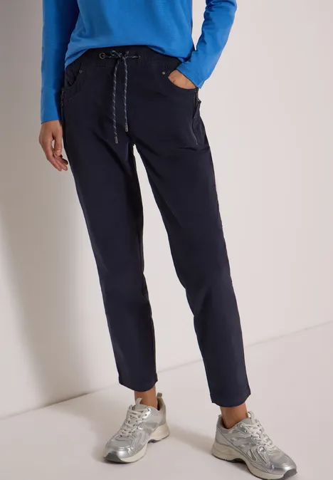 Femme en pantalon bleu marine décontracté à cordon et baskets argentées sur fond clair.