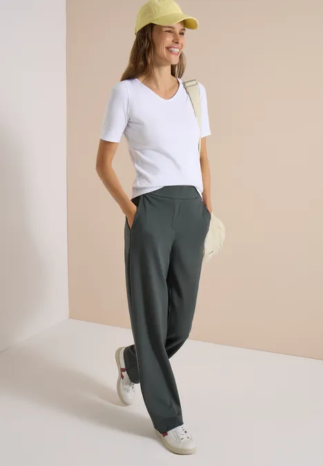 Jeune femme en tee-shirt blanc, pantalon large vert, baskets blanches et casquette jaune devant un mur beige.