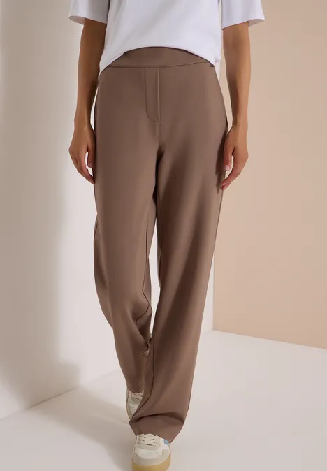Une personne porte un pantalon ample marron et des baskets blanches devant un mur sobre.
