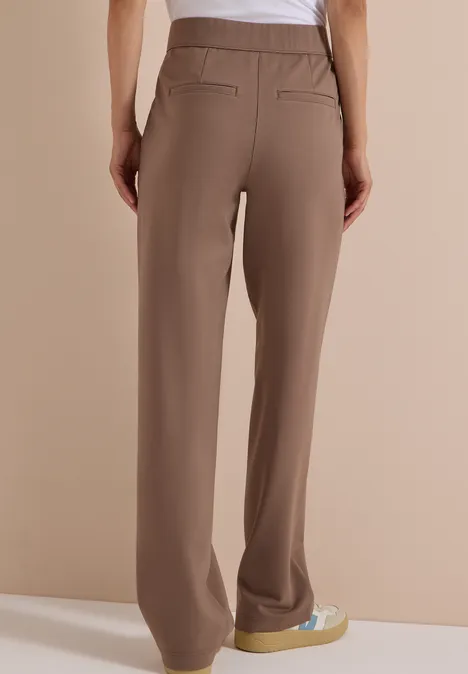 Femme vue de dos portant un pantalon fluide beige à poches et baskets compensées sur fond neutre.