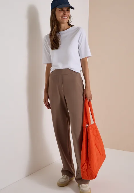 Une femme porte un t-shirt blanc, un pantalon marron, une casquette bleue et un grand sac orange.