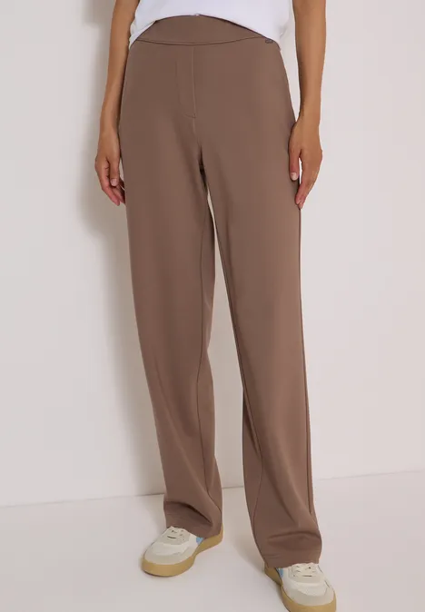 Personne en pantalon marron ample coupe droite et baskets blanches sur fond clair.
