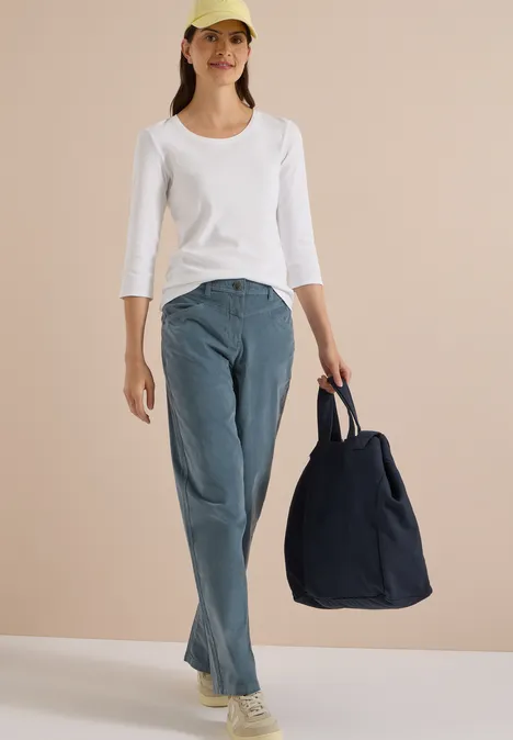 Femme en t-shirt blanc à manches longues, jean et casquette, tenant un grand sac bleu marine sur fond beige.