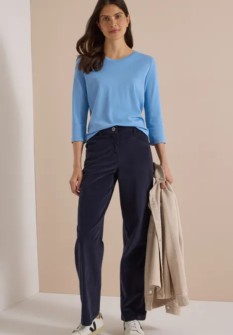 Femme en haut bleu clair et pantalon bleu marine, tient un manteau beige et pose devant un fond neutre.