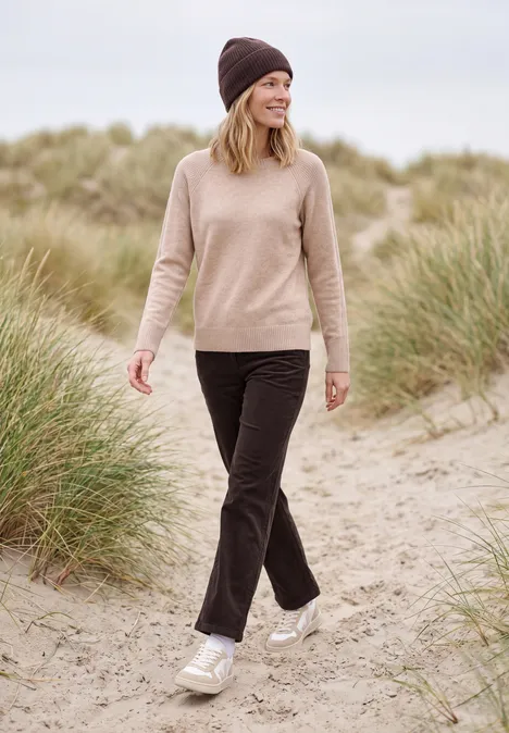 Eine Frau mit Wollmütze und beigem Pullover spaziert in Sneakern über sandige Dünen.