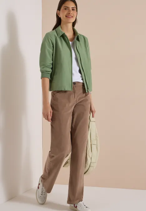 Une femme porte une veste verte, un t-shirt blanc, un pantalon marron et tient un sac beige.